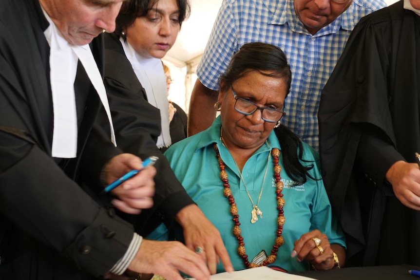 Yamatji woman Glenda Jackamarra signs documents surrounded by legal officials wearing black and white robes.
