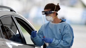 A woman in personal protective equipment holds a swab as she tests a person in their car for coronavirus.