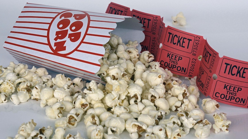 A packet of popcorn laying on its side with popcorn spilled on a table and movie tickets in the background