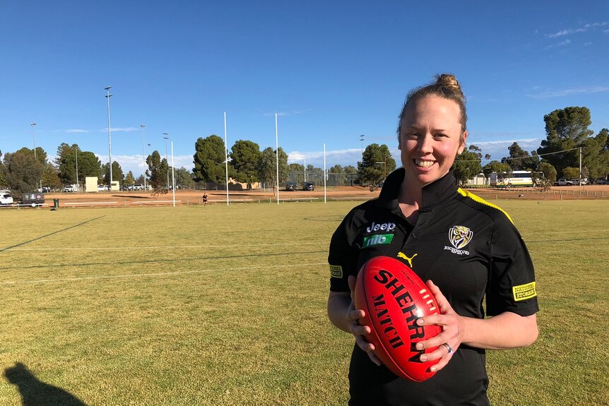 Woman in Richmond Football Club t-shirt standing on a country football oval holding a football.