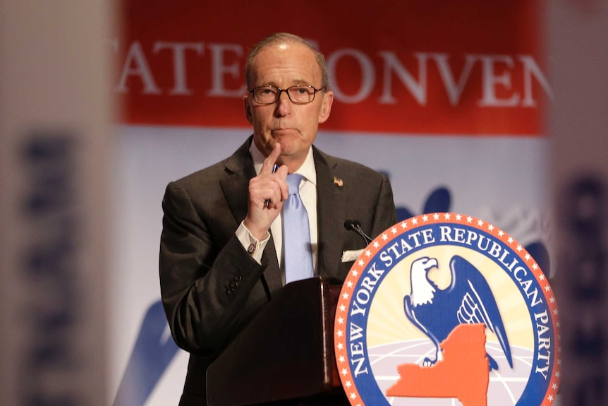 Larry Kudlow speaking, with a pen in his right hand and behind the sign of "New York State Republican Party"