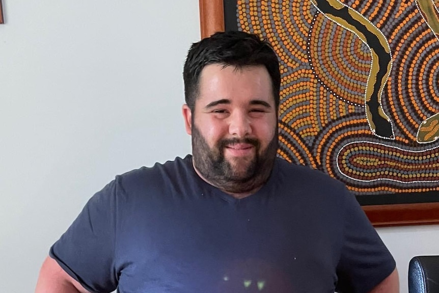 A heavy set man with a beard smiles in front of a indigenous art painting