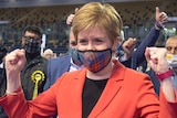 Wearing a red jacket and tartan facemask, Nicola Sturgeon clenches her fists in a victory gesture in front of supporters