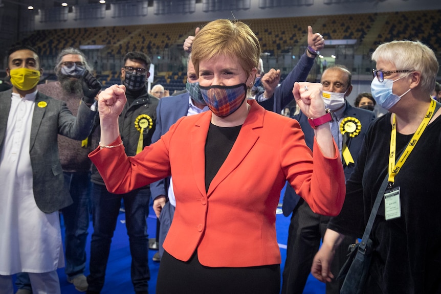 Wearing a red jacket and tartan facemask, Nicola Sturgeon clenches her fists in a victory gesture in front of supporters