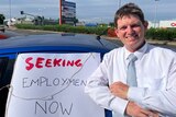 A middle aged man in a white office shirt leans against a blue car with a sign that says, ' Seeking employment now'. 