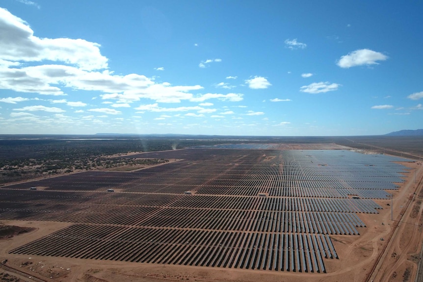 A solar power plant on land with red soil and trees in the distance.