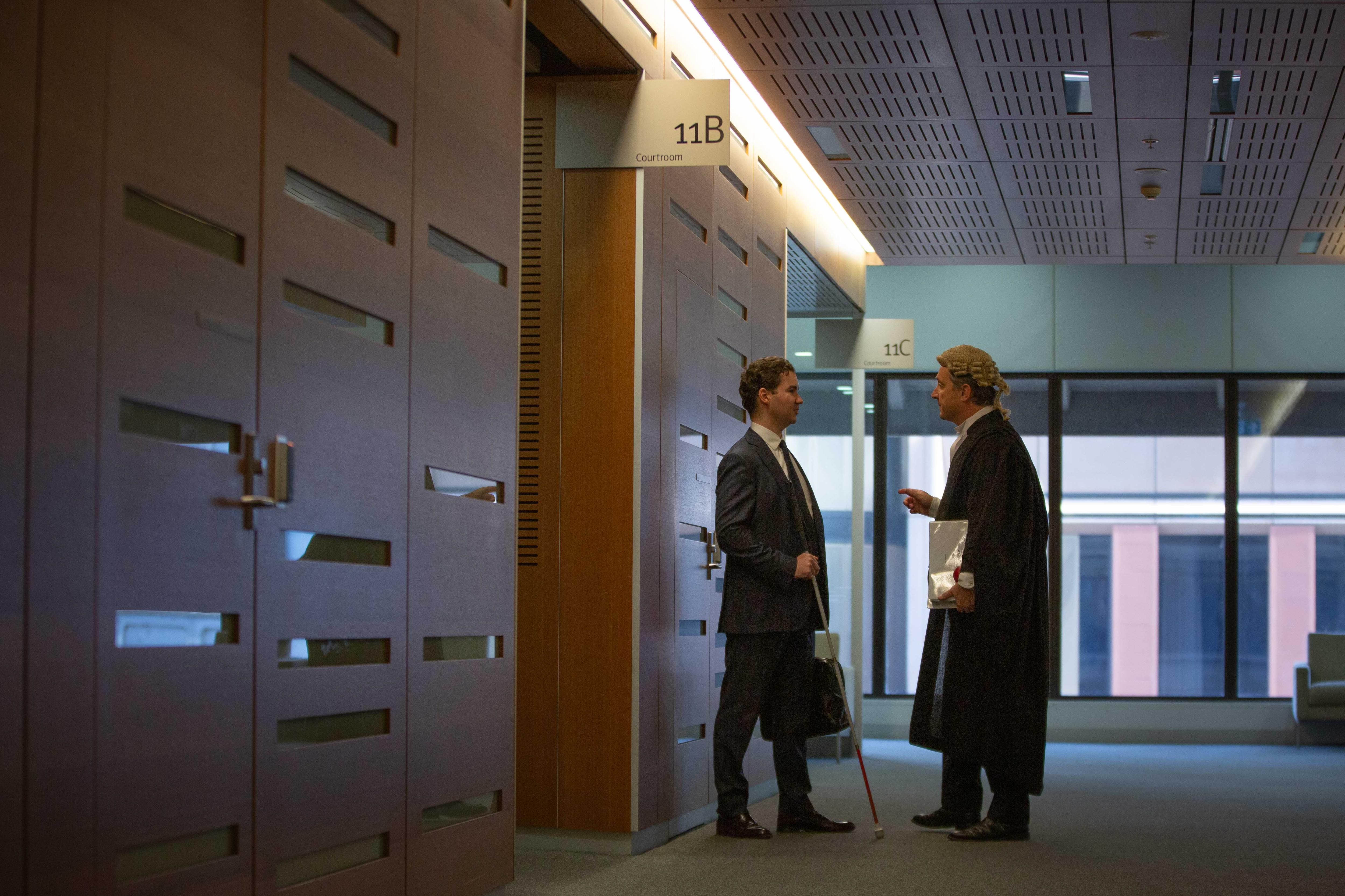 A white man with short hair, blue suit and white cane talking with a barrister in a hallway