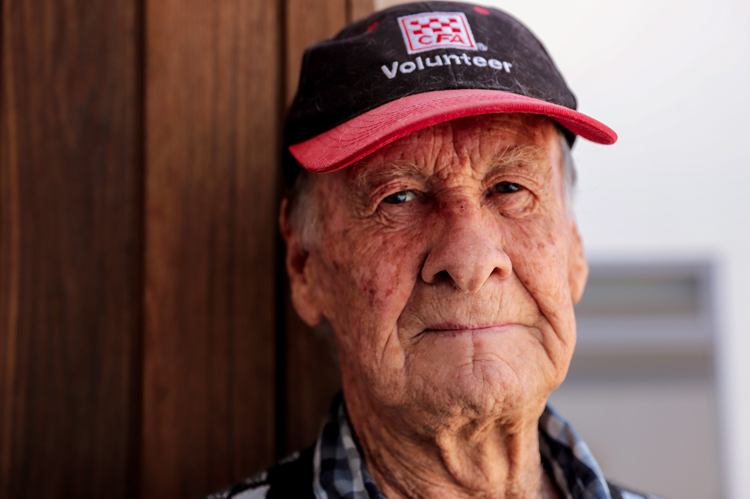 Older man stares wearing CFA Volunteer hat with his back to a wooden wall