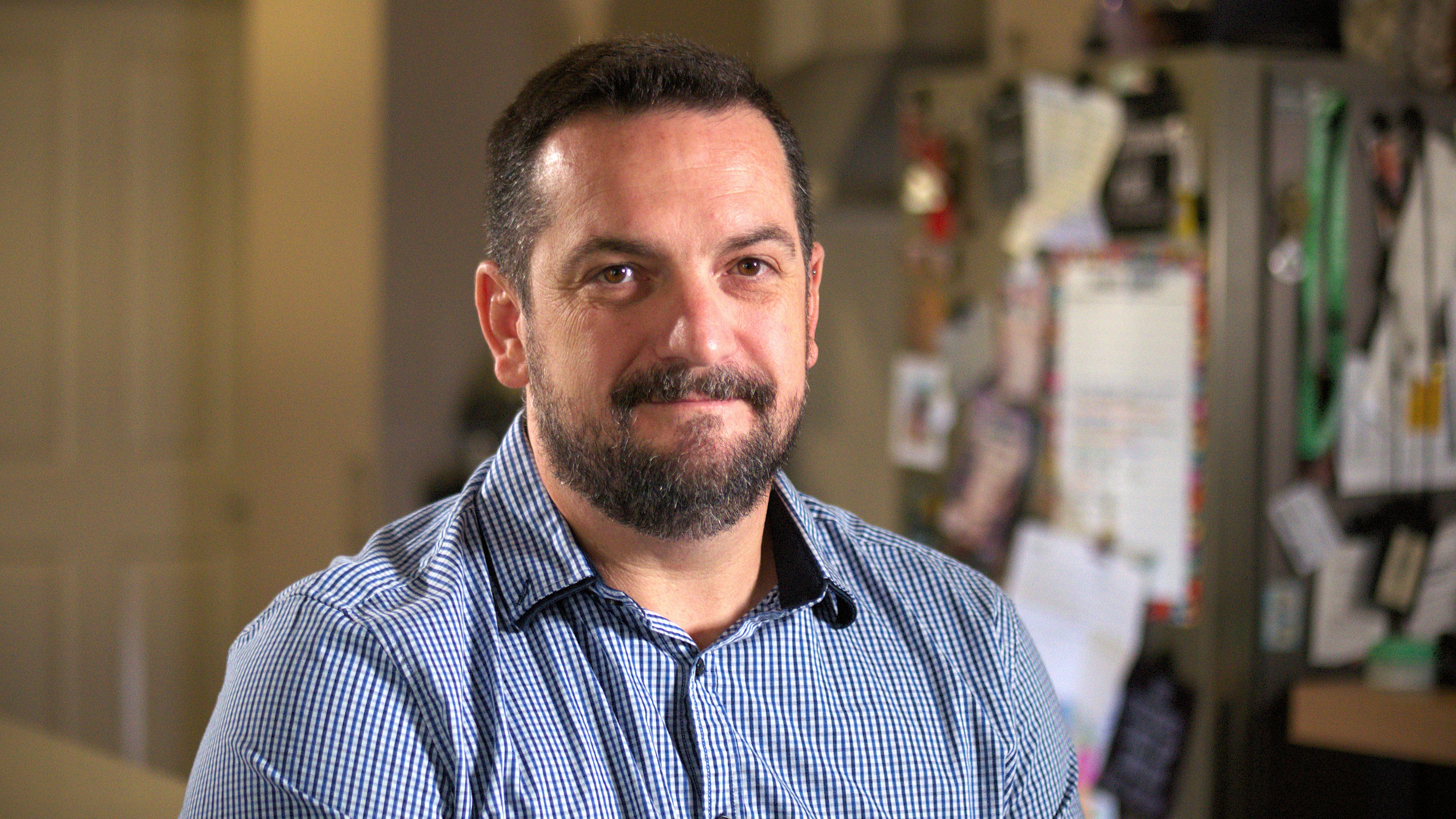 A man in a blue button up looks reflectively at the camera.