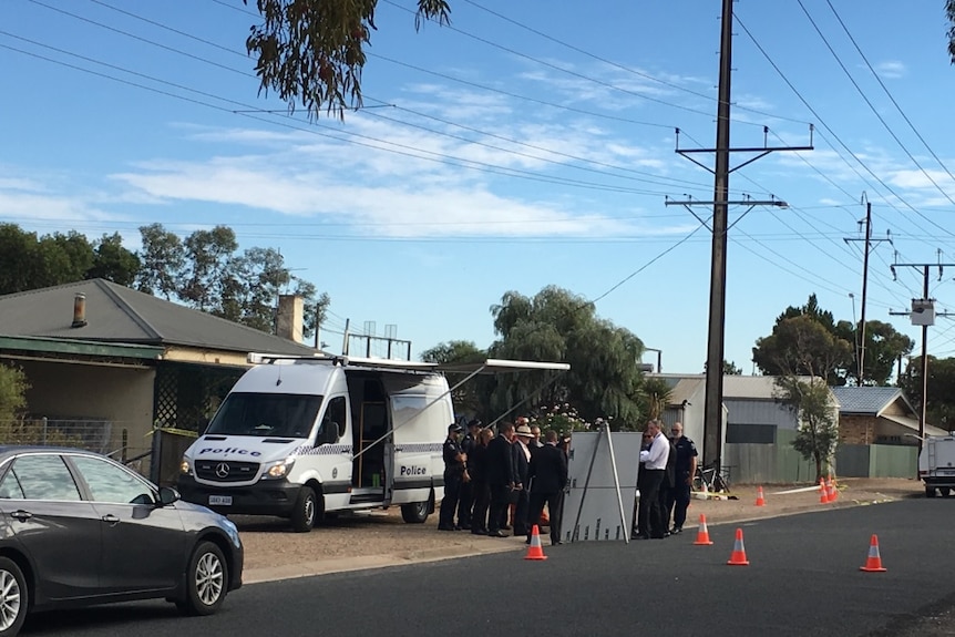 A white van and police detectives outside a house, with cones across the roadway.