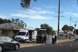 A white van and police detectives outside a house, with cones across the roadway.