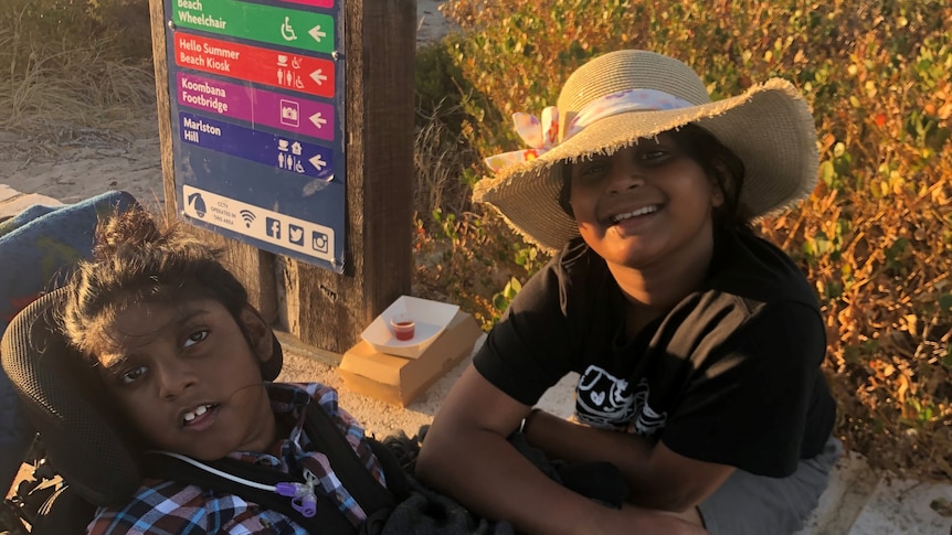 A little boy sits in a wheelchair near a sign at the beach with his sister 