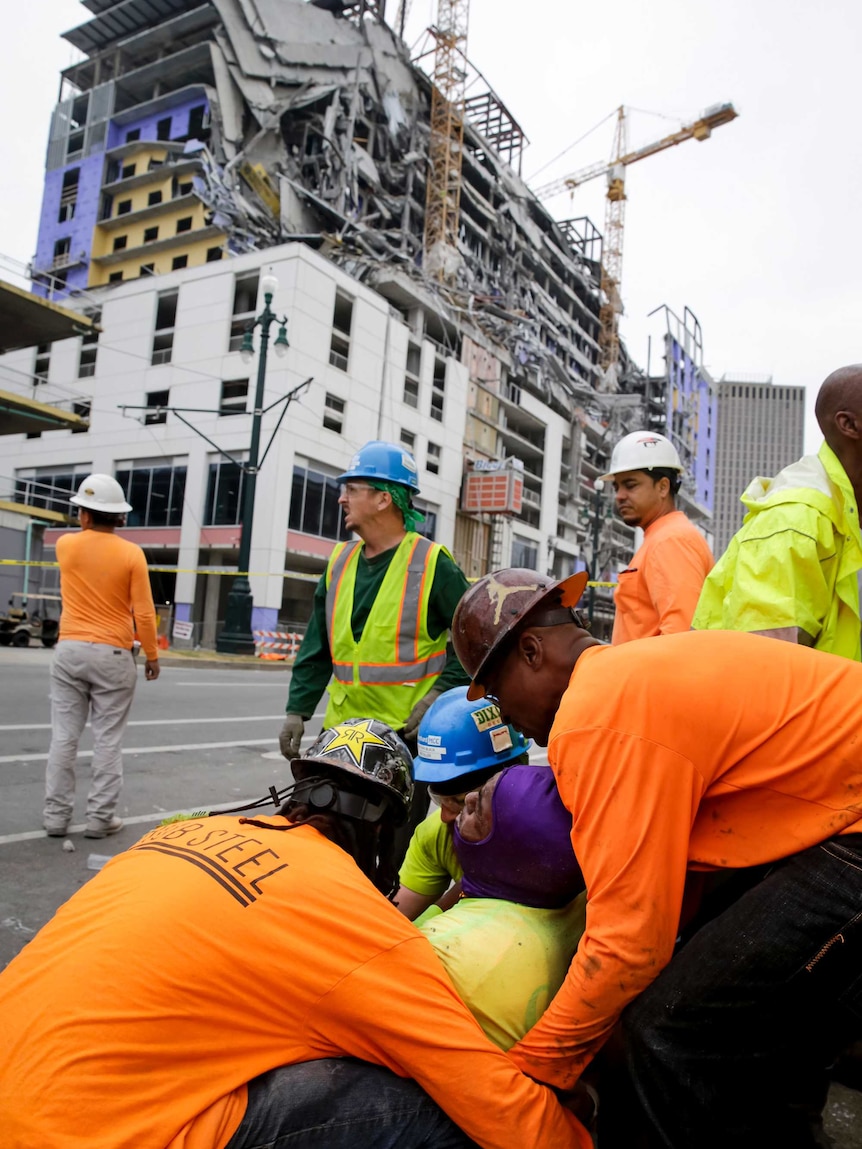 Three construction workers hold up an injured man on the side of the road with a collapsed building in the background