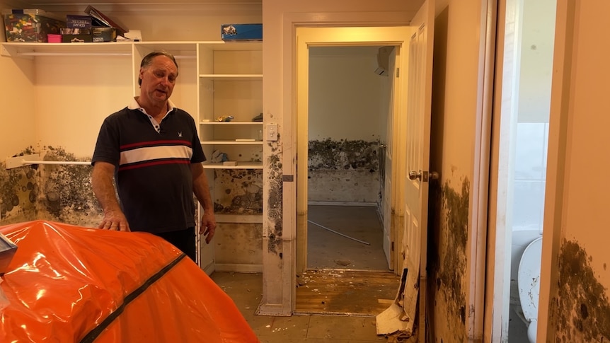 A man in a dark shirt stands in his mouldy bedroom.