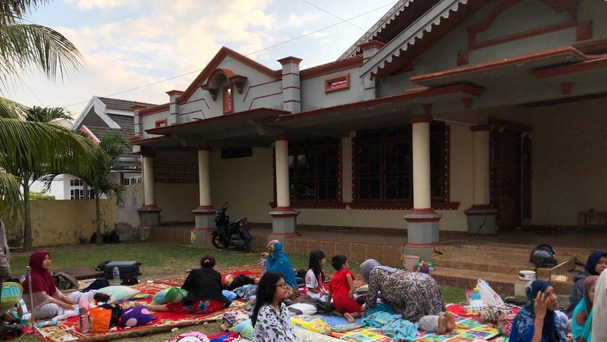 Women and children sleeping and sitting down on mats in a garden in front of a house with a crack running through it