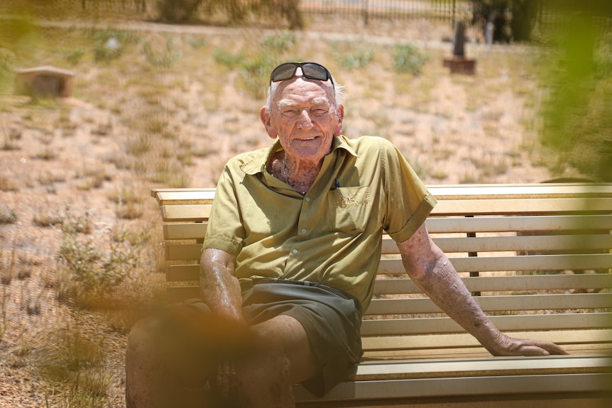 An elderly man sits on a bench in a cemetery, smiling at the camera.