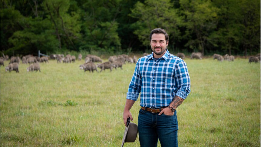 Photo of a man with a checkered shirt holding an akubra standing out in the field.