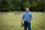Photo of a man with a checkered shirt holding an akubra standing out in the field.