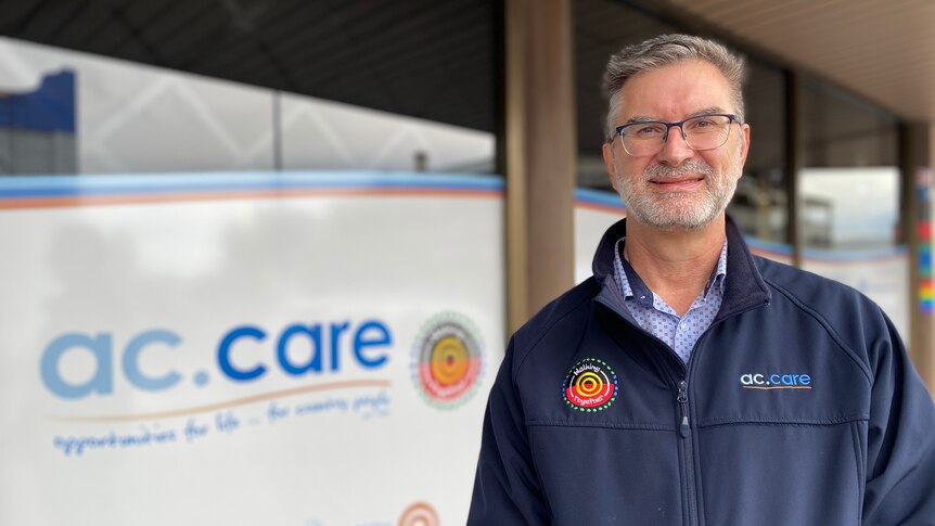 A man with glasses smiles at the camera with a sign behind him reading 'a c care'