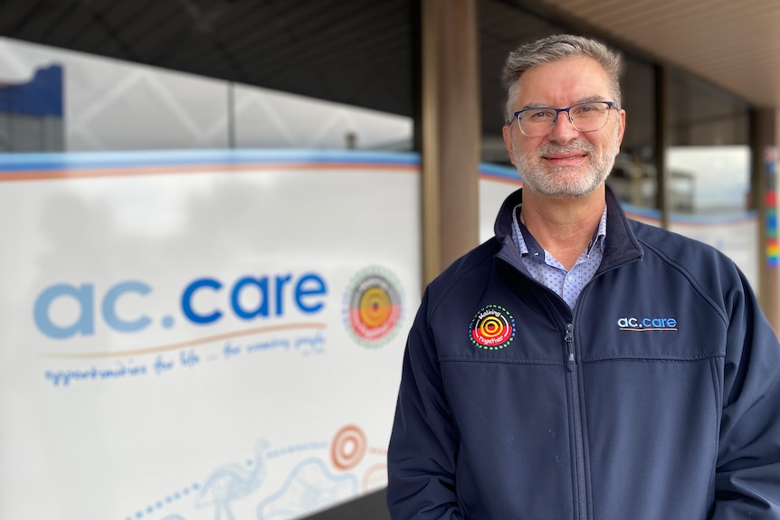 A man with glasses smiles at the camera with a sign behind him reading 'ac care'
