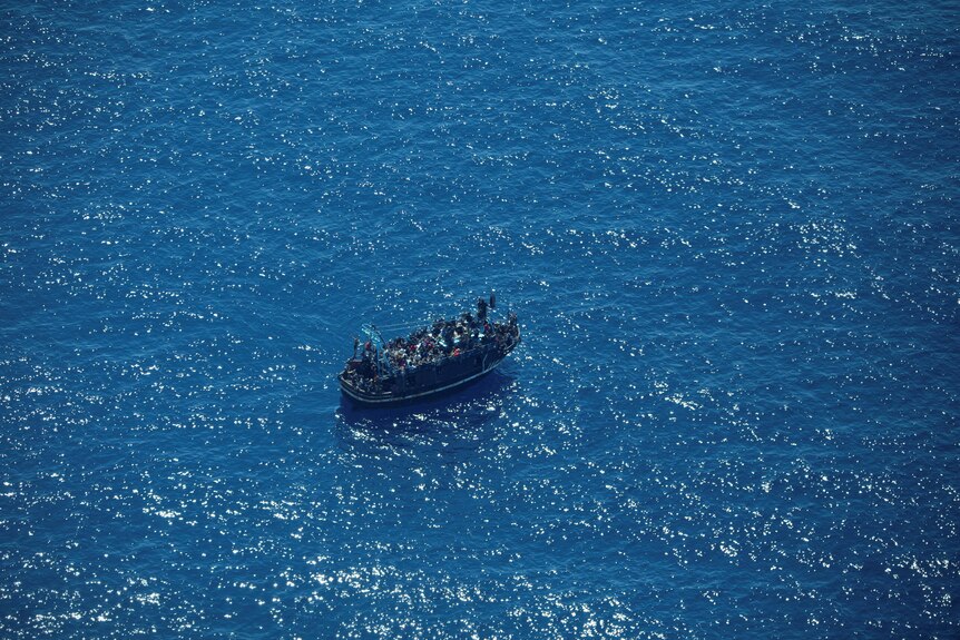 A boat packed full of people is seen from above on a blue sea