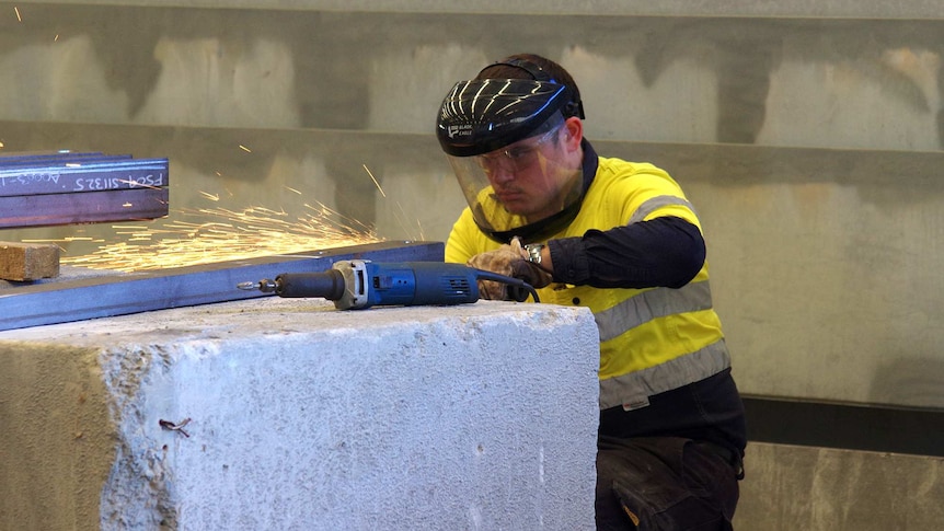 A man wearing a protective mask grinds metal on a concrete block at a manufacturing facility.