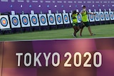 Two people walk in front of archery targets with the sign 'Tokyo 2020' in the foreground.
