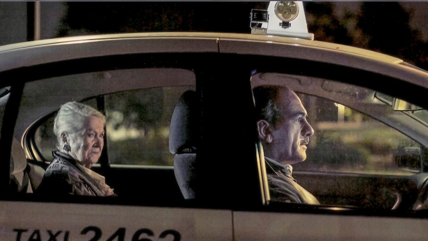 A Middle-Eastern taxi driver at the wheel with an elderly white woman in the back