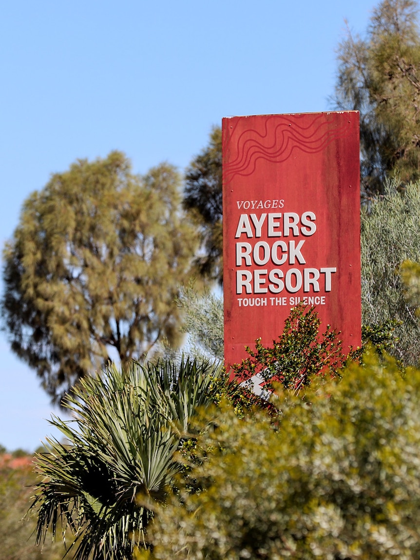 A red sign reading Ayers Rock Resort is visible between bush and trees beneath a blue sky