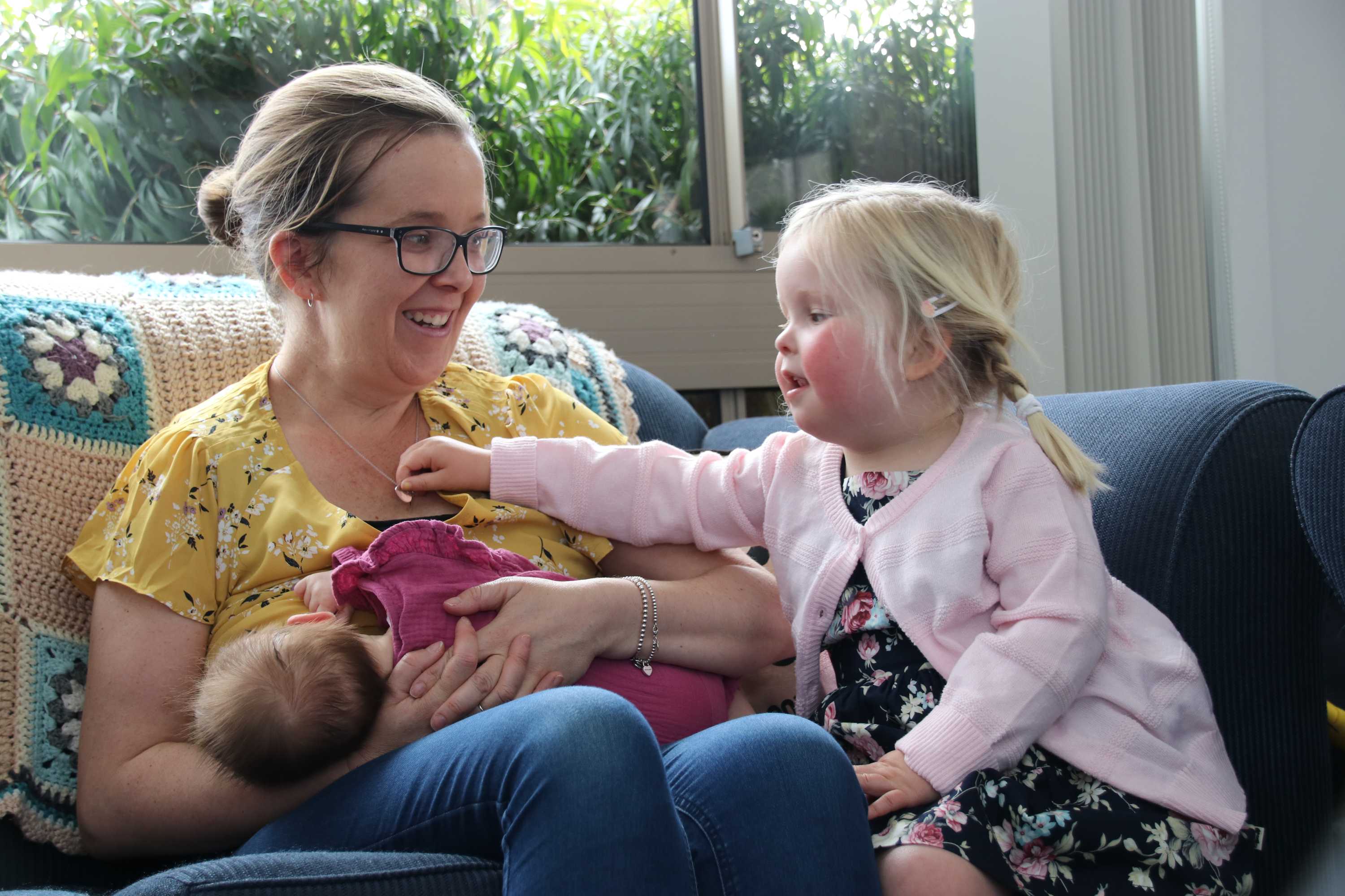 A toddler is reaching over to hold her mother's necklace, as the mother breastfeeds her other daughter. The mother is smiling.