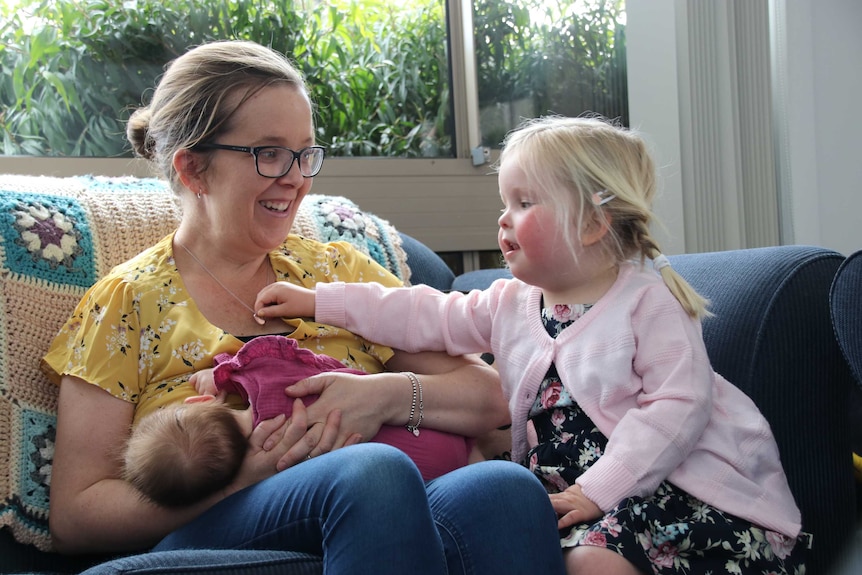 A toddler is reaching over to hold her mother's necklace, as the mother breastfeeds her other daughter. The mother is smiling.