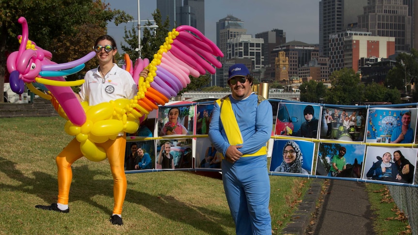 A man wearing a bird made out of balloons stands by a city river next to a man wearing photo-covered wings.