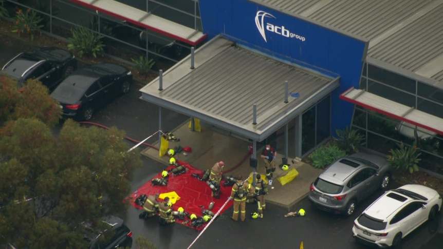 Firefighters kneel and walk around equipment laid out on a red tarp outside a building saying ACB Group.