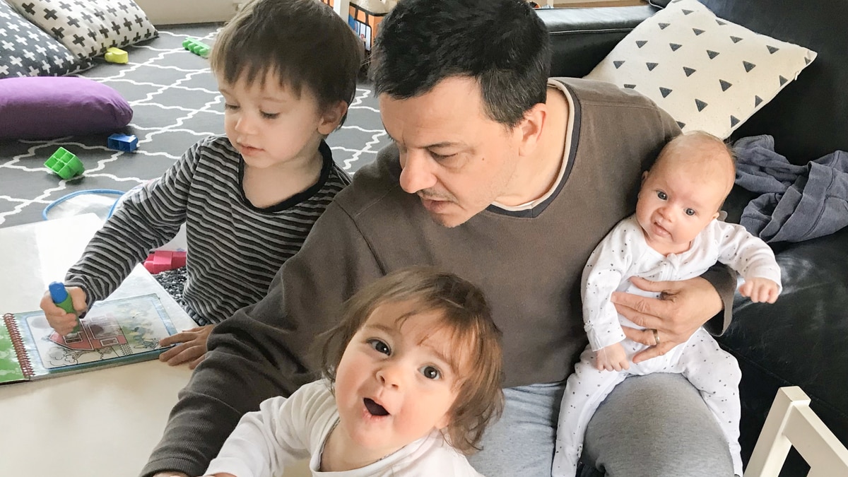 Mark Tamhane plays with two of his young children at a table while juggling a baby on his knee.