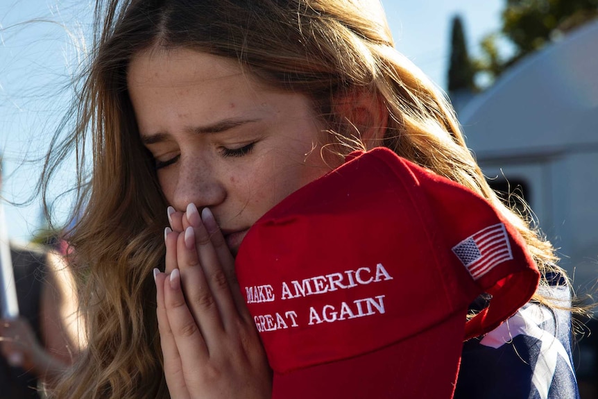 A young girl puts her hands together in prayer while holding a "Make America Great Again" cap in support of Donald Trump.