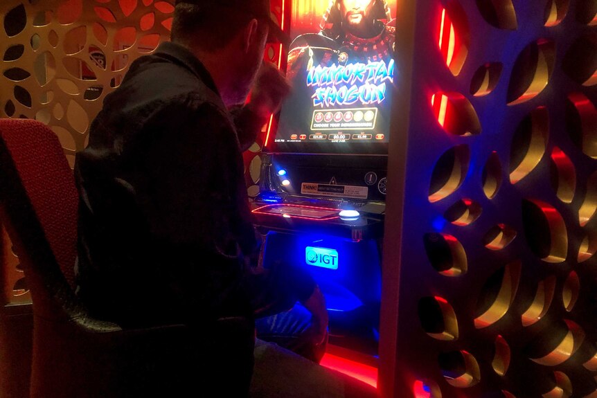 Man wearing black shirt and cap sits backed turned to camera at neon pokie machine.