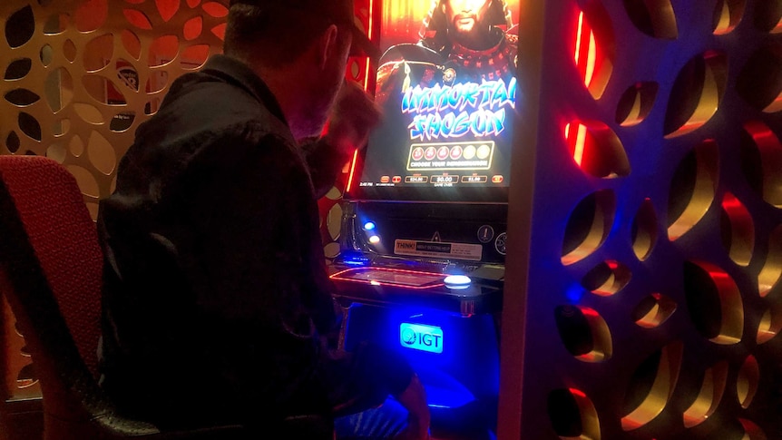 Man wearing black shirt and cap sits backed turned to camera at neon pokie machine.