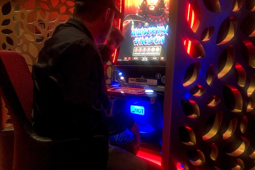 Man wearing black shirt and cap sits backed turned to camera at neon pokie machine.