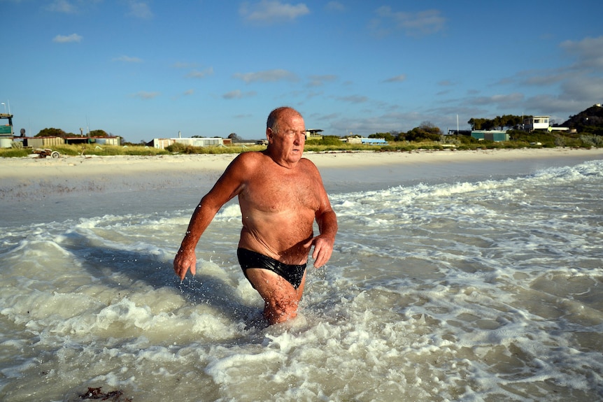 A tanned older man in budgie smugglers strides into the surf