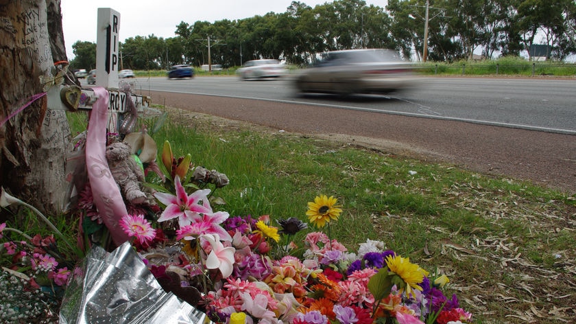 Groundshot of cross with RIP and lots of flowers next to roadside as cars whizz by