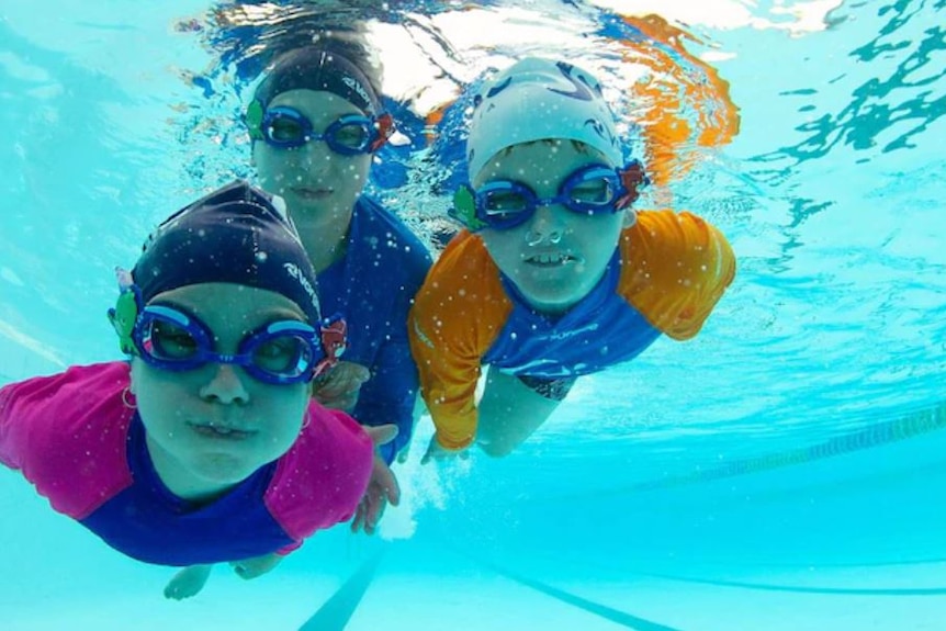 Three kids swim towards the camera below the surface of a pool, lanes visible behind them. They wear goggles and swimming caps.