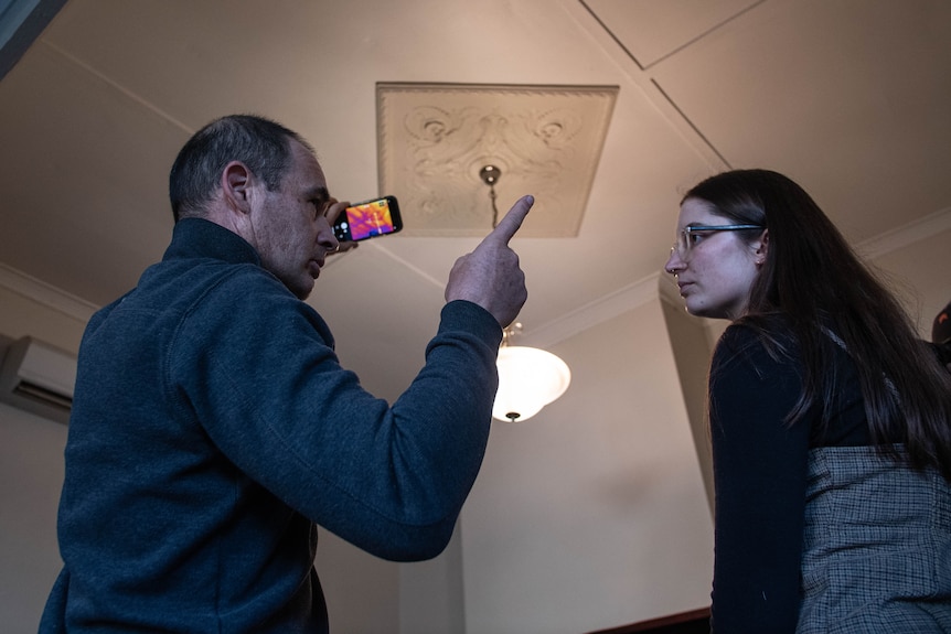 A middle-aged man in jumper points to ceiling while holding a phone with thermal imaging app towards ceiling, talking to woman