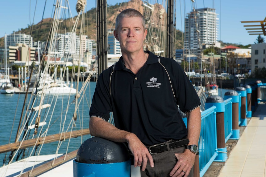 A man with short hair stands at a marina, with boats in the background.