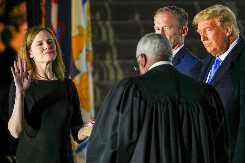 Amy Coney Barrett holding up her left hand to Clarence Thomas while Donald Trump watches on
