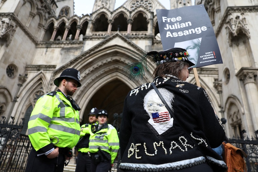 Several police officers surround a woman outside a large old building. The woman is holding a sign saying "Free Julian Assange"