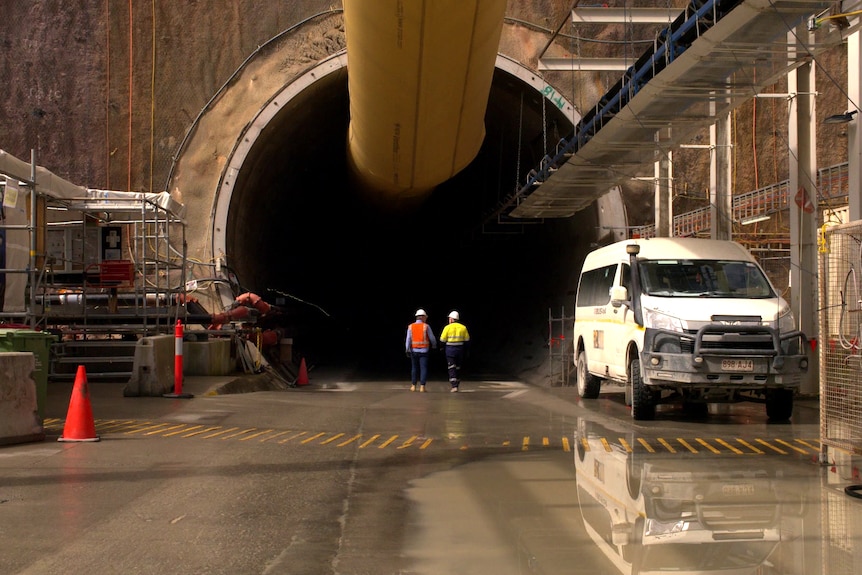 Two men wearing hard hats and hi-visibility walk into the opening of a large tunnel, several times their height.