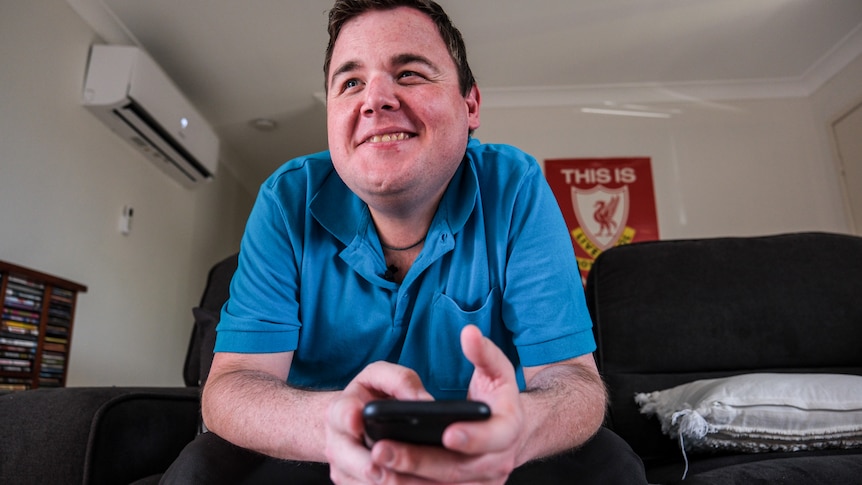 Conor Smith sits on a couch at home, dressed in a bright blue shirt. He's leaning forward while holding his black iPhone.