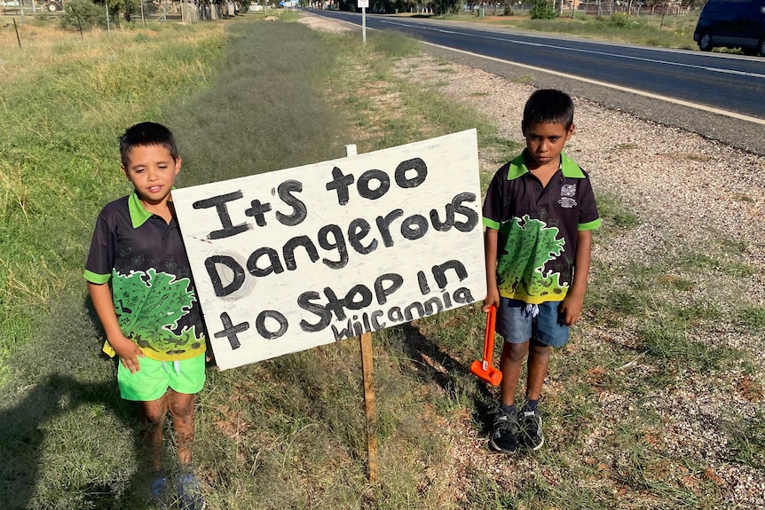 Two young Aboriginal boys stand either side of a sign reading 'It's too dangerous to stop in Wilcannia'.