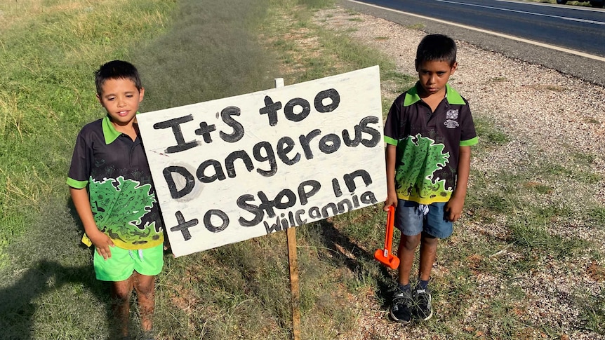 Two young Aboriginal boys stand either side of a sign reading 'It's too dangerous to stop in Wilcannia'.