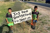 Two young Aboriginal boys stand either side of a sign reading 'It's too dangerous to stop in Wilcannia'.